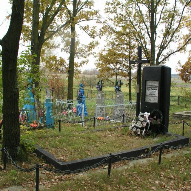 Tombe des Polonais assassinés par l'Armée insurrectionnelle ukrainienne en 1943, située dans un cimetière avec des arbres et un monument noir avec des inscriptions. Des couronnes et des fleurs sont disposées autour.