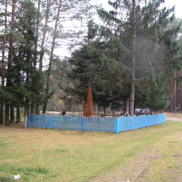 A memorial site in Rudnia Łęczyńska with a blue wooden fence surrounding a grave marked by an obelisk. Tall pine trees grow around the site.