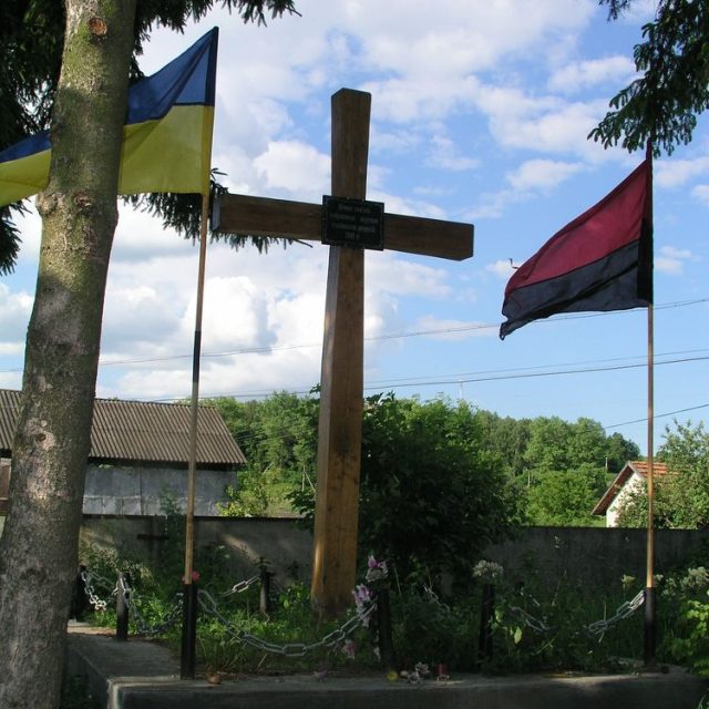 Wooden cross on a grave with Ukrainian flags on both sides. The grave is surrounded by a chain fence and flowers. Inscription in Ukrainian commemorates victims of Stalinist repressions in 1941.