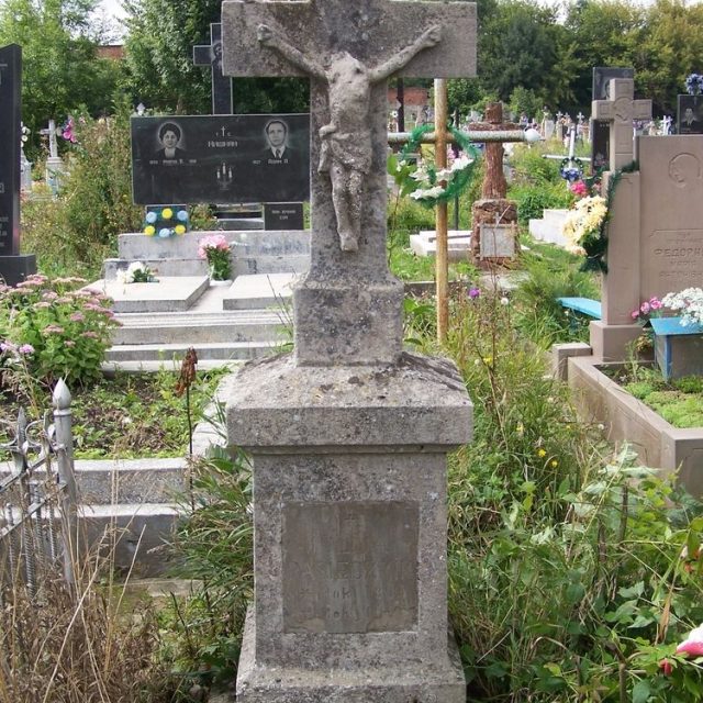 Gravestone of Julia Pasiecznik with a cross on a two-tiered pedestal. The cross features a relief of Christ and an inscription panel on the front. The pedestal is topped with a four-sided roof.