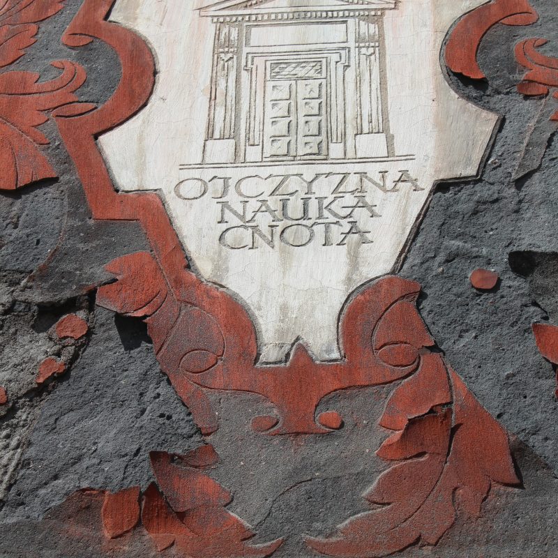 Napis 'OJCZYZNA CNOTA NAUKA' na ścianie Kościoła św. Katarzyny w Wilnie, otoczony dekoracyjnymi elementami w kolorze czerwonym i szarym.