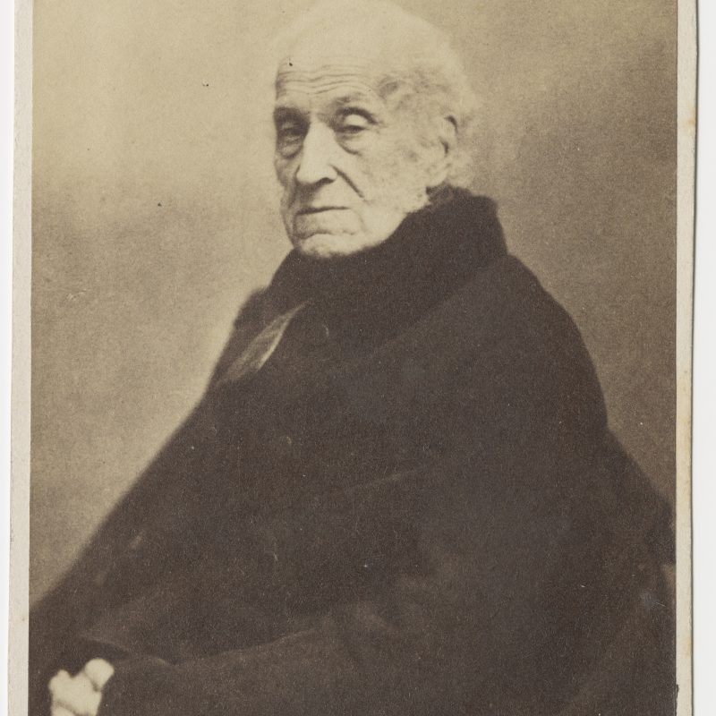 Sepia-toned photograph of an elderly man with white hair, wearing a dark coat, seated and looking towards the camera.