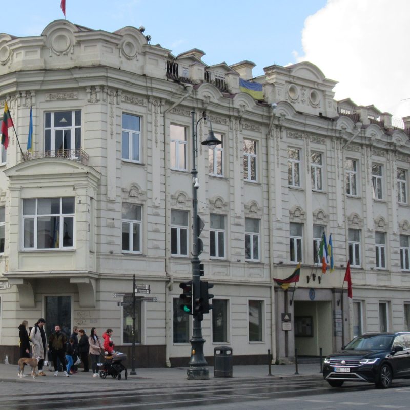 Historic building in Vilnius, corner of Mickiewicza and Arsenalska streets, formerly housing the cafe 'U Rudnickiego'. The facade features ornate decorations and multiple flags. Pedestrians and a car are visible.