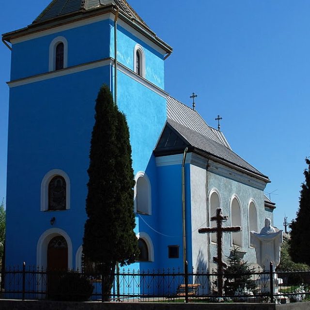L'ancienne chapelle de la Visitation de la Sainte Vierge Marie à Mościska, avec une façade bleue et une tour. Entourée d'arbres et d'une clôture métallique, elle présente des fenêtres cintrées et une croix devant.