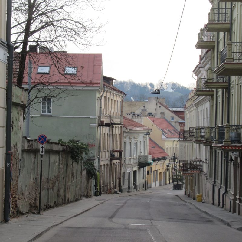Une rue de Vilnius bordée de part et d'autre de bâtiments historiques, avec des balcons et un trottoir étroit. La rue est en pente, on aperçoit un panneau d'interdiction de stationnement. À l'arrière-plan, des arbres et des collines.