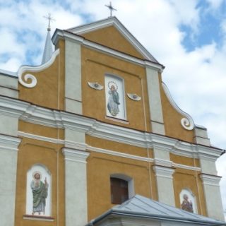 Fasada kościoła parafialnego pw. Wszystkich Świętych w Miżyńcu z trzema malowidłami religijnymi i trójkątnym szczytem na tle pochmurnego nieba.