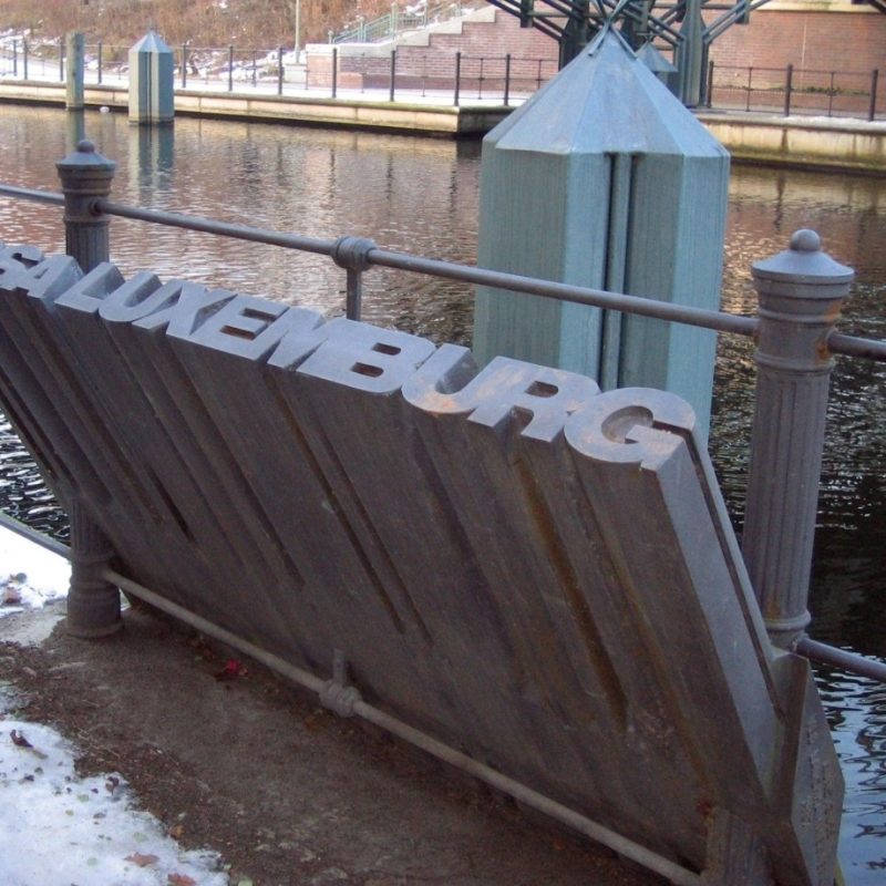 Des lettres métalliques formant l'inscription « Rosa Luxemburg » sur une surface en pierre près du Landwehrkanal à Berlin, avec une balustrade métallique et l'eau en arrière-plan.