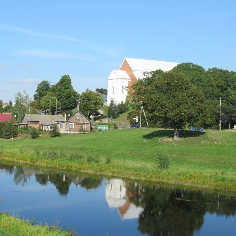St. George's Church in Kėdainiai, a Gothic basilica with a high roof and polygonal apse, surrounded by trees and reflected in a nearby river, with rural houses in the foreground.