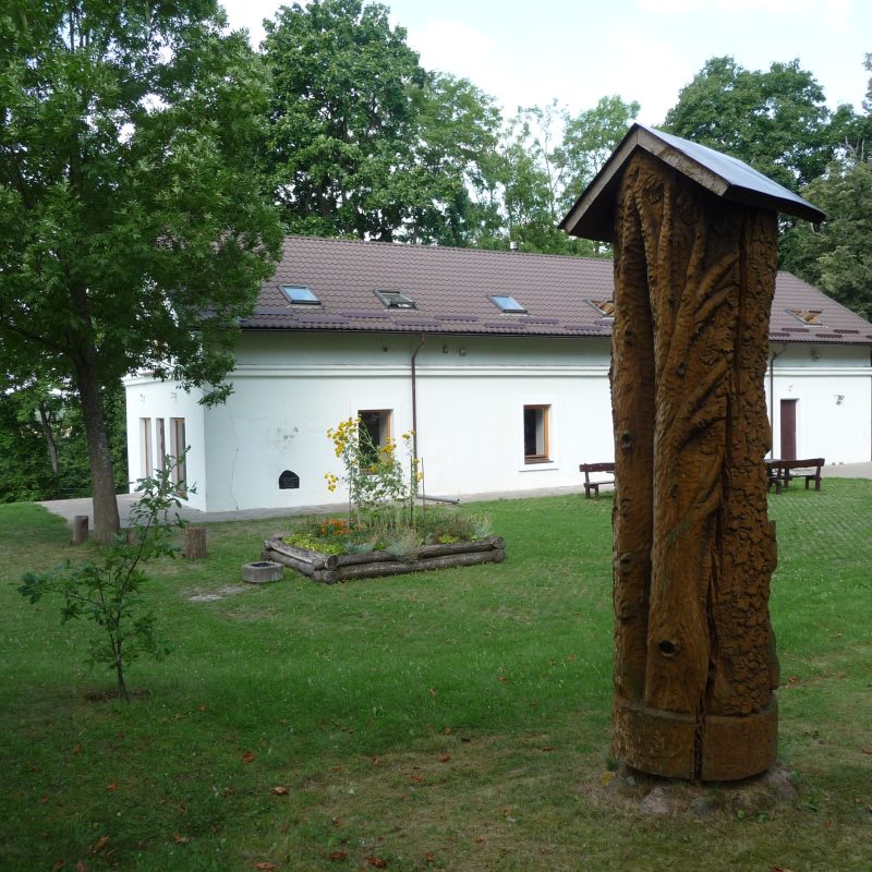 Un ancien grenier restauré à Szetejnie, entouré de verdure. Au premier plan, une sculpture en bois aux ornements raffinés trône sur la pelouse.