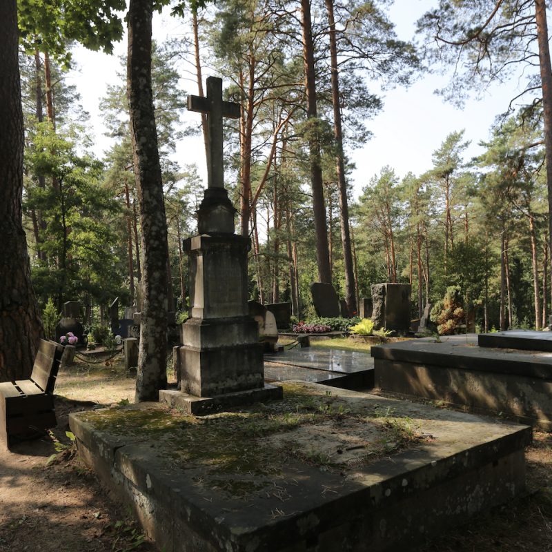 Kamienny krzyż na trzystopniowym cokole z wyrytą tablicą, otoczony płytami z piaskowca na betonowym fundamencie, na zalesionym cmentarzu.