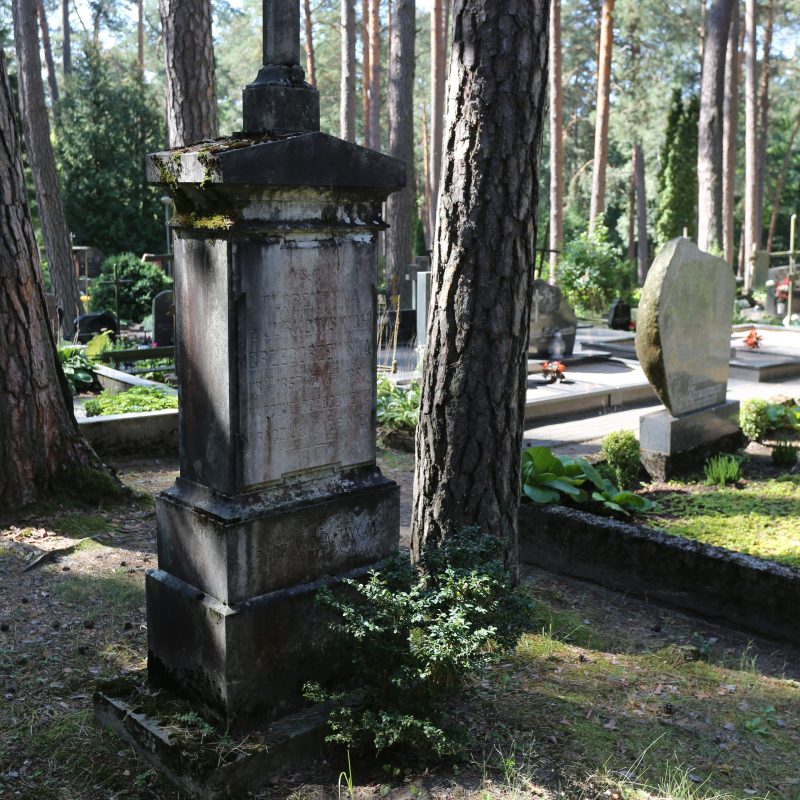 Nagrobek Florentyny Grzegorzewskiej z białym wapiennym krzyżem na trzystopniowym cokole. Widoczna inskrypcja na pierwszym stopniu. Otoczony drzewami na cmentarzu.
