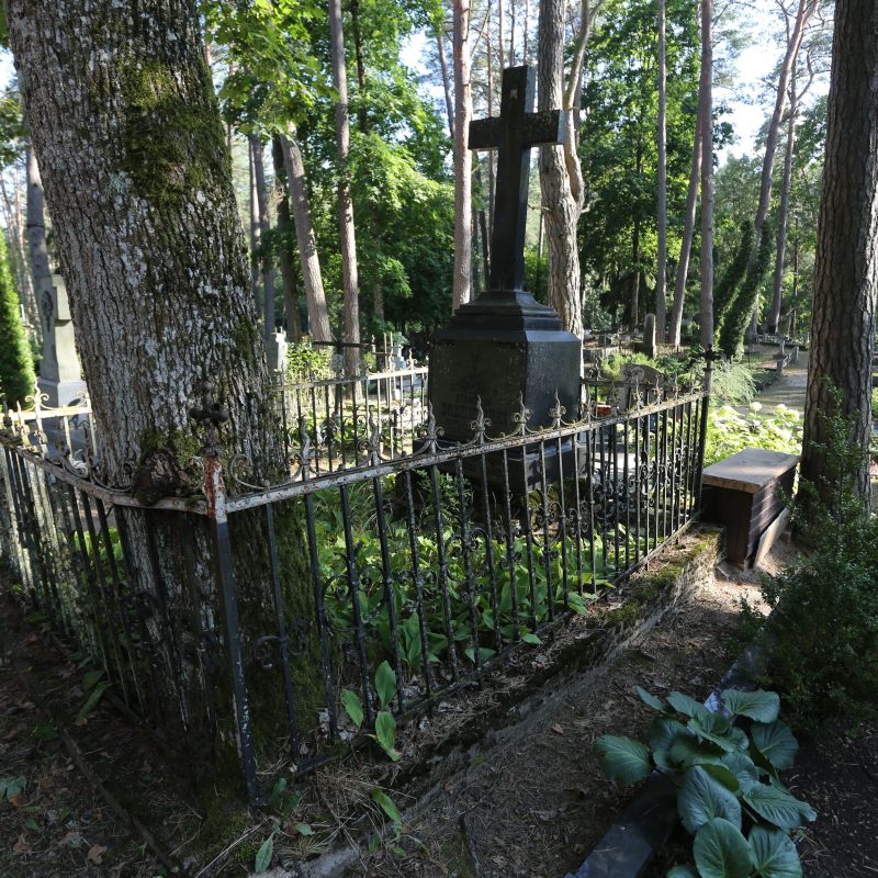 Croix en granit noir sur un piédestal à deux niveaux entourée d'une clôture en fer sur une base en béton. La tombe est dans un cimetière boisé avec des arbres et de la verdure.