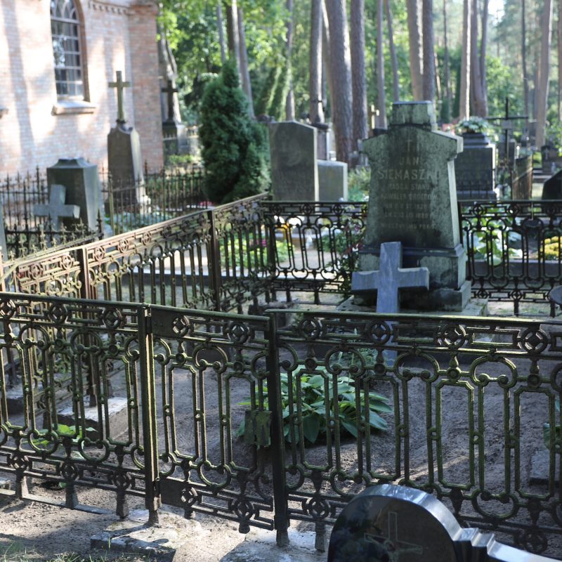 Tombe de Jan Siemaszko avec un piédestal en granit à deux niveaux et une croix en granit gris cassée. La tombe est entourée d'une clôture en fer incomplète sur des blocs de béton.
