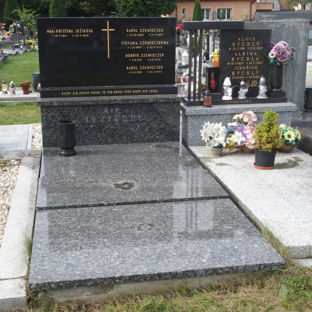 Pierre tombale des familles Ježík et Szewieczek avec une croix et des inscriptions. La dalle en pierre a un vase. Des fleurs et une petite plante sont placées devant.
