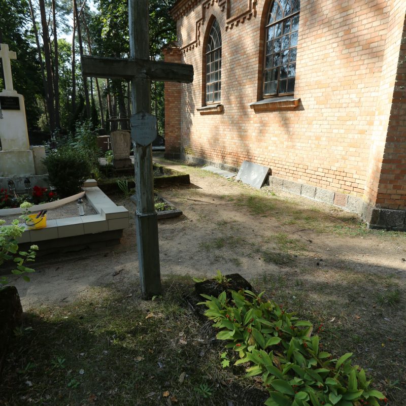 Дерев'яний хрест із залишками срібної фарби та цинковою табличкою у формі гербового щита. Попереду бетонна облямівка та рослини. На задньому плані цегляна будівля з арковими вікнами.
