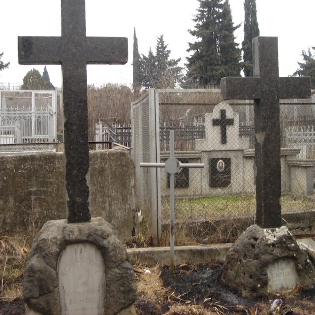 Dwa czarne kamienne krzyże na nieregularnych podstawach na cmentarzu. Lewy krzyż ma marmurową płytę epitafijną z inskrypcją. W tle widoczny metalowy płot i inne groby.