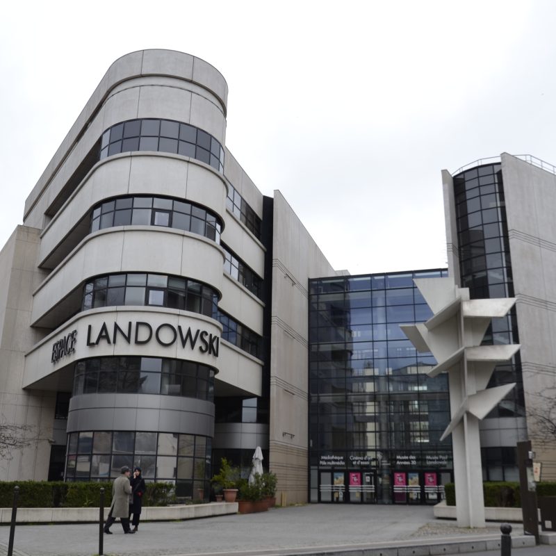 Будівля Espace Landowski з сучасною архітектурою, великими скляними вікнами та помітною вивіскою 'Landowski'. Перед входом стоїть скульптура.