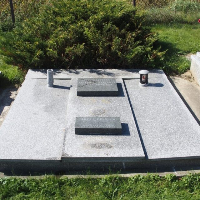 Grób oznaczony płytą nagrobną z dwiema rytowanymi tablicami i mniejszą metalową tabliczką. Płyta nagrobna składa się z czterech mniejszych płyt, otoczona trawą i innymi grobami.