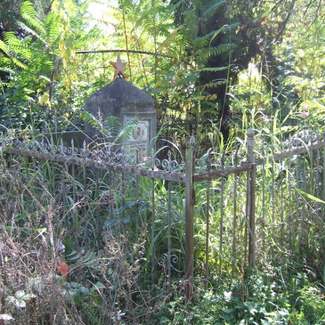 Obélisque en ciment de la tombe de Jerzy Sokołowski avec une étoile rouge au sommet, entouré d'une clôture métallique. Une plaque en marbre avec une inscription en russe et une photo du défunt est visible. La zone est envahie par la végétation.