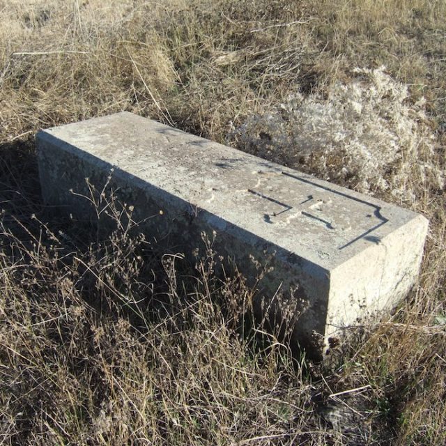 Pierre tombale rectangulaire aux bords biseautés, inscrite en russe, posée sur un champ herbeux.