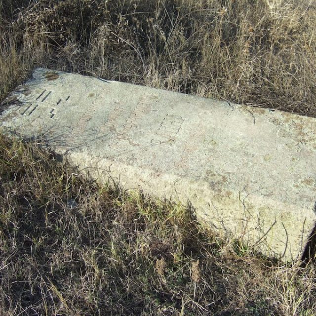 Pierre tombale d'Anna Wacka, dalle rectangulaire aux bords biseautés, partiellement enterrée, avec une inscription en russe.