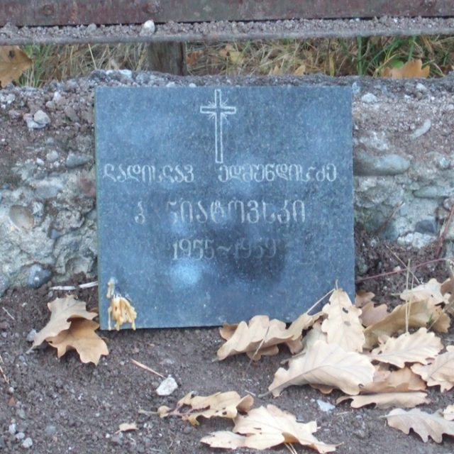 Pierre tombale en marbre noir avec une croix gravée et des inscriptions dans un script étranger, entourée de feuilles tombées et de terre.