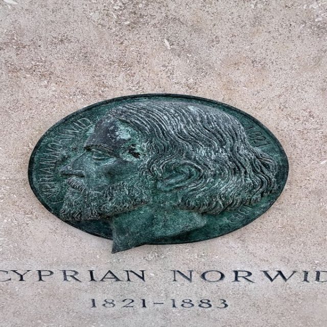 Nagrobek Cypriana Kamila Norwida na cmentarzu w Montmorency. Zawiera brązowy relief profilu Norwida i wyryty napis 'Cyprian Norwid 1821-1883' na kamiennej powierzchni.