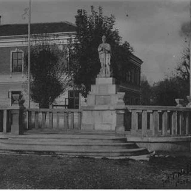 Photographie historique d'un monument à Lviv, représentant un homme sur un piédestal entouré d'une balustrade. À l'arrière-plan, un bâtiment avec des fenêtres et des arbres.