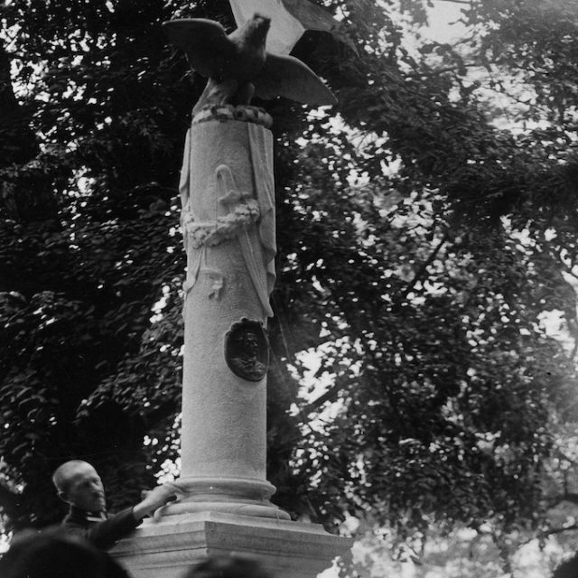 Monument to Tadeusz Kosciuszko in Zhovkva, column with Kosciuszko medallion and eagle sculpture on top. Trees visible in the background.