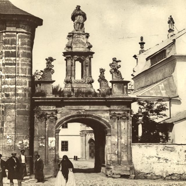 Archiwalna fotografia barokowej Bramy Katedralnej w Kamieńcu Podolskim, z rzeźbą św. Jana Nepomucena na szczycie. Przy bramie widoczna jest grupa przechodniów.