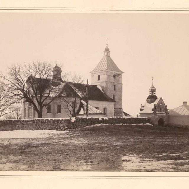 Photographie historique de l'église de l'Assomption à Rockat, montrant l'église avec une grande tour, entourée d'arbres sans feuilles et d'un mur de pierre, prise avant l'incendie de 1898.