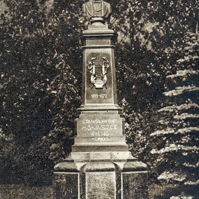 Historic photograph of the monument to Stanislaw Moniuszko in Vilnius, with bust on pedestal with inscriptions and decorative elements, surrounded by trees.