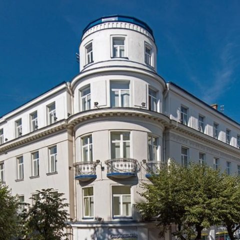 Trzypiętrowy budynek z zaokrąglonym narożnikiem i uskokową wieżą, z dużymi oknami i dekoracyjną fasadą. Drzewa częściowo zasłaniają dolną część budynku.