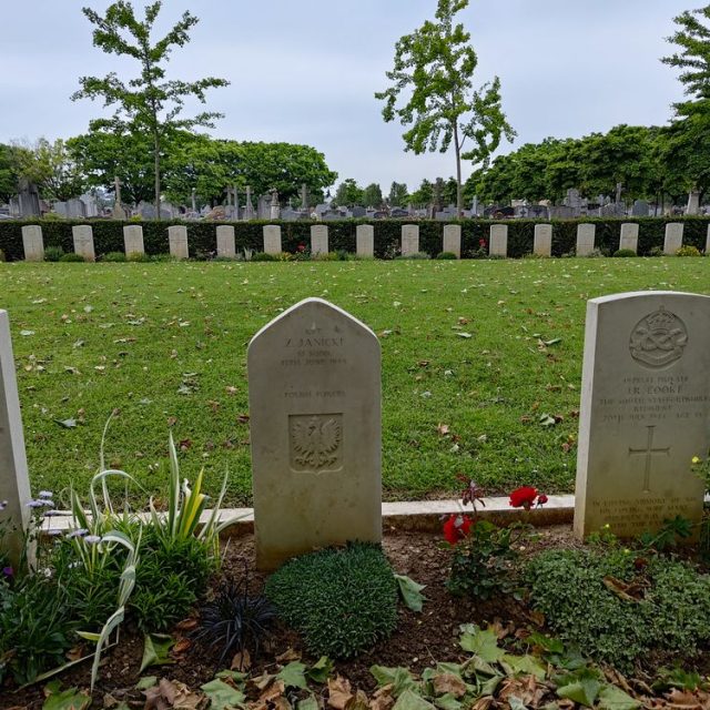 Grave of Captain Zbigniew Janicki at the Western Cemetery in Le Mans, marked by a headstone with an eagle emblem and surrounded by grass and flowers. Adjacent graves of other soldiers are visible.
