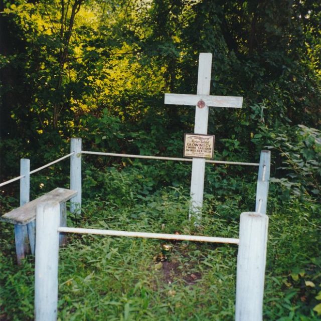 Biały krzyż z tabliczką stoi na trawiastym terenie otoczonym prostym drewnianym ogrodzeniem, oznaczając grób polskich ofiar UPA w zalesionej okolicy.