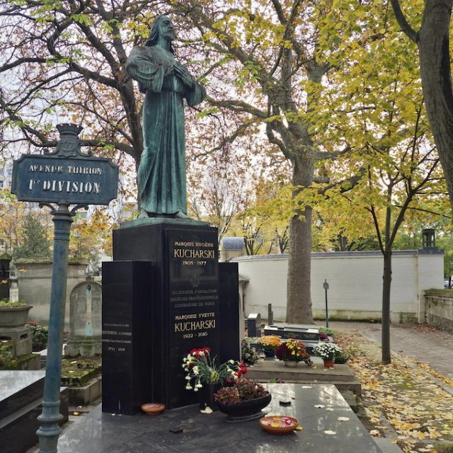 Grave of Aleksander, Eugeniusz, and Yvette Kucharski with a bronze statue of Christ in a long robe, hands on chest. Black marble slab with inscriptions, surrounded by trees with autumn leaves.