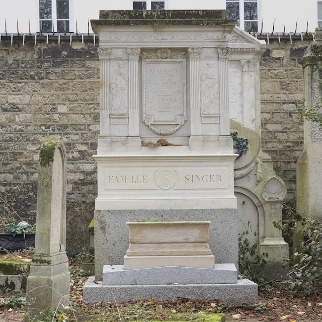 Tombe de la famille Singer avec deux figures allégoriques dans des niches. À gauche, une femme en robes antiques avec un enfant, représentant 'La Charité'. À droite, une femme avec des attributs comme un caducée et une ancre, représentant 'L’Industrie'.