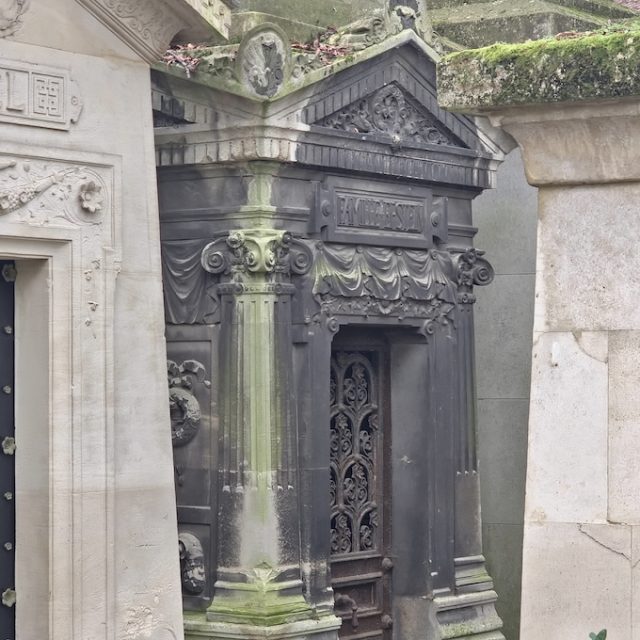 Mausoleum with a gabled roof, tympanums with a frieze, and Ionic pilasters. Draped material decorates the exterior. The entrance has wrought iron doors with floral motifs. Inside, a stained glass window and inscribed plaques are visible.