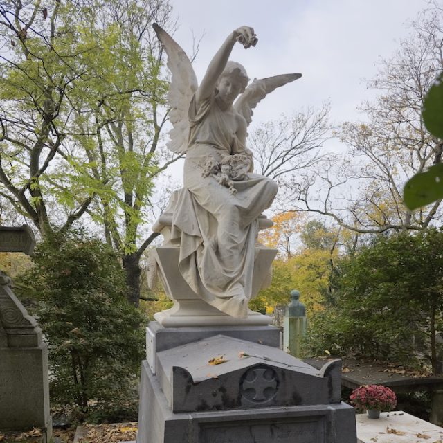 A marble tomb with a seated angel holding flowers. The tomb features a cross in a circle on the front. Trees with autumn leaves surround the scene.