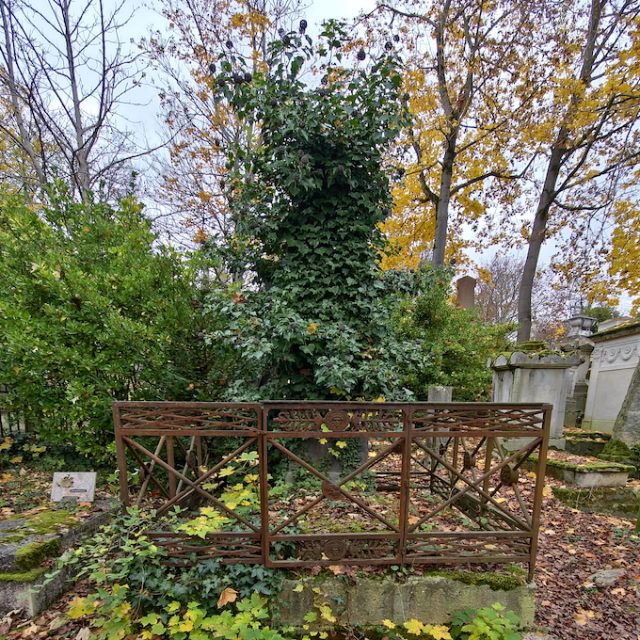 Grave of the Roguet family and Henryk Steinkeller with an obelisk covered in ivy, surrounded by an iron fence with palm leaf motifs. Autumn trees and other tombstones in the background.