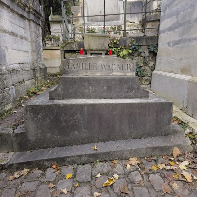 Tombe de la famille Wagner avec un socle en gradins et l'inscription gravée 'FAMILLE WAGNER'. Une jardinière en pierre avec des bougies rouges est placée à la tête. Des feuilles tombées couvrent le sol pavé.