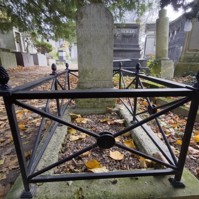 Tombe de Sophie Wołowska avec une stèle sur un piédestal, sommet semi-circulaire, entourée d'une clôture en fer. Des feuilles tombées couvrent le sol. La tombe a été restaurée en 2010.