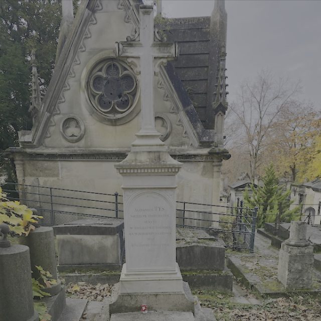 Gravestone of Aleksander Tys with a tall cross on a rectangular pedestal, featuring an inscription. Behind, a stone structure with a circular window and triangular roof. Autumn trees and other graves in the background.
