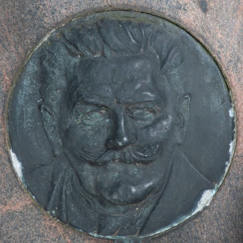 Bronze medallion with a relief portrait of a man with slicked-back hair, prominent eyebrows, sunken eyes, small nose, and mustache. He wears a jacket, shirt, and tie.