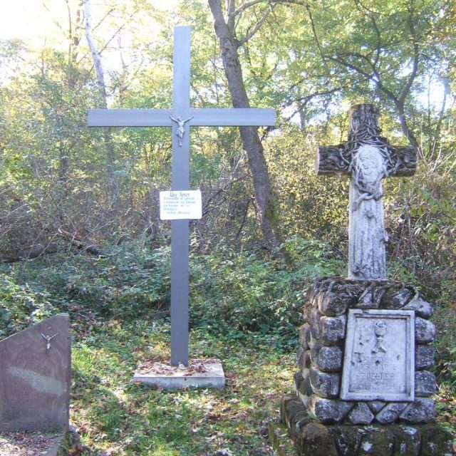 Metal cross with a plaque in Ukrainian and a stone cross with a relief in a wooded area, symbolizing the graves of UPA victims in Puźniki.