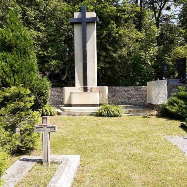 Centralny pomnik krzyża w kwaterze żołnierzy polskich na Cmentarzu Janowskim, otoczony zielenią i mniejszymi nagrobkami.