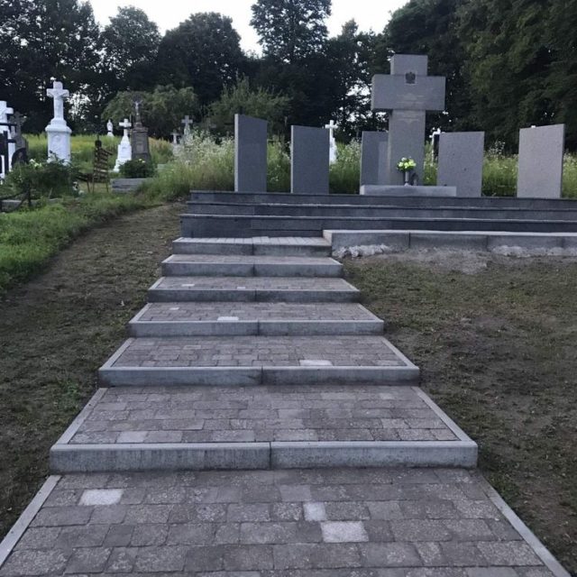 Kamienne schody prowadzą do miejsca pamięci z centralnym krzyżem i pionowymi tablicami otoczonymi zielenią. Miejsce upamiętnia polskie ofiary masakry z 1944 roku w Palikrowach.