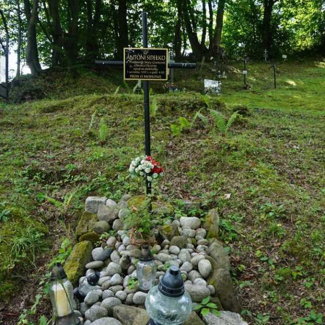 Grób Antoniego Sidełko, polskiego strażnika granicznego, na starym cmentarzu w Mniszku. Grób oznaczony metalowym krzyżem z tabliczką, otoczony kamieniami i zniczami.