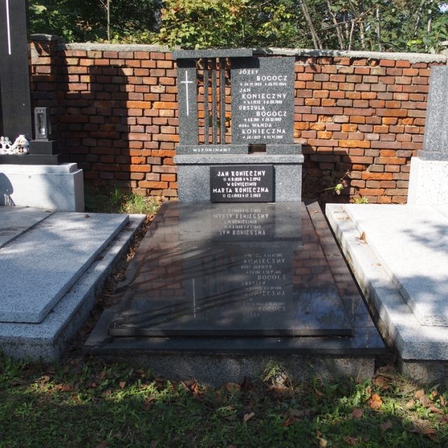 Nagrobek rodziny Bogocz i Koniecznych z inskrypcjami na wysokiej podstawie i krzyżem. Otoczony murem z cegieł i sąsiednimi grobami. Kwiaty i świece w pobliżu.