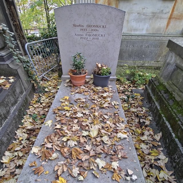 Grave of Anna and Marcin Olomucki with a flat, stepped slab covered in fallen leaves. At the head, a rectangular stele with an arched top and two potted plants.